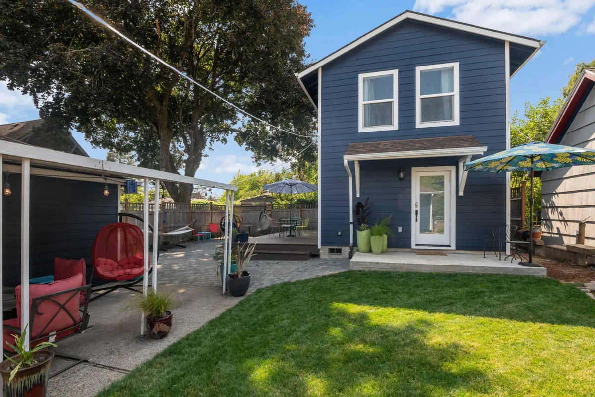 A cheerful exterior view of a blue house with a lush green lawn and a welcoming outdoor seating area, complete with colorful decorative umbrellas. The scene exudes a relaxed atmosphere, suggesting opportunities for outdoor gatherings, morning coffee, and unwinding in the garden.