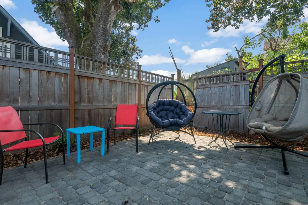 This exterior image showcases a charming stone patio with a hanging chair, blue side table, and vibrant red chairs. The patio is surrounded by greenery and exudes a relaxed vibe, ideal for enjoying coffee, reading, or socializing outdoors.