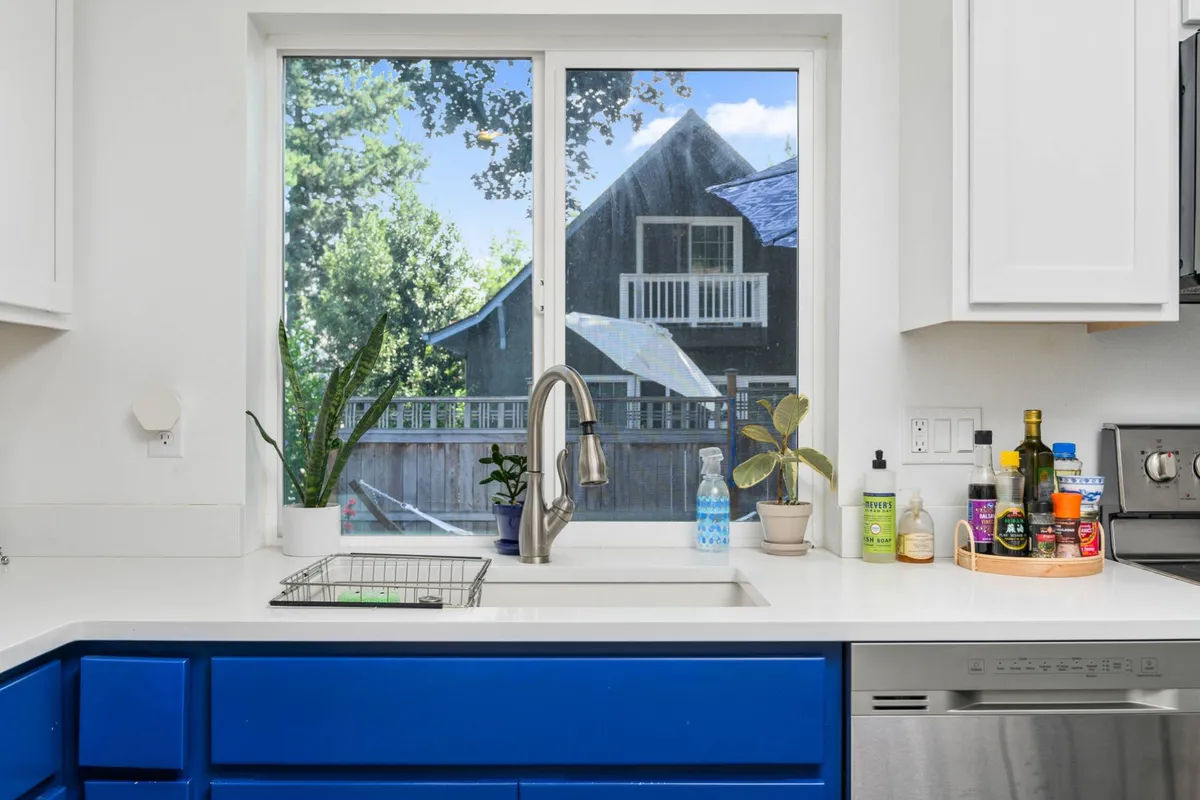 A bright, cheerful kitchen with blue cabinets, potted plants, and a window offering leafy views. The setup, filled with natural light, feels both inviting and energizing—ideal for a morning coffee before work or a break during a busy day.