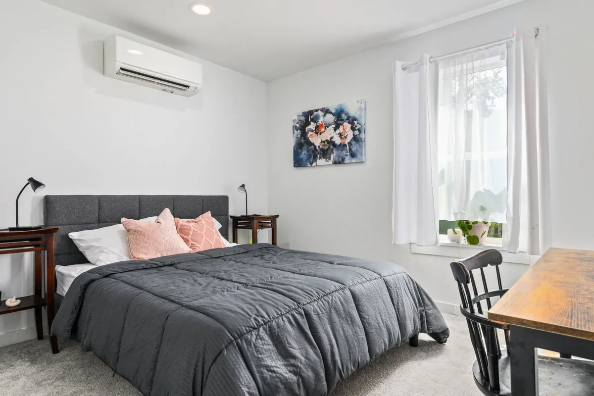 This image features a serene bedroom with a plush bed, stylish artwork above the headboard, and a desk positioned by the window. The space is thoughtfully decorated to evoke calm and comfort, with ample natural light pouring in to create a welcoming atmosphere.