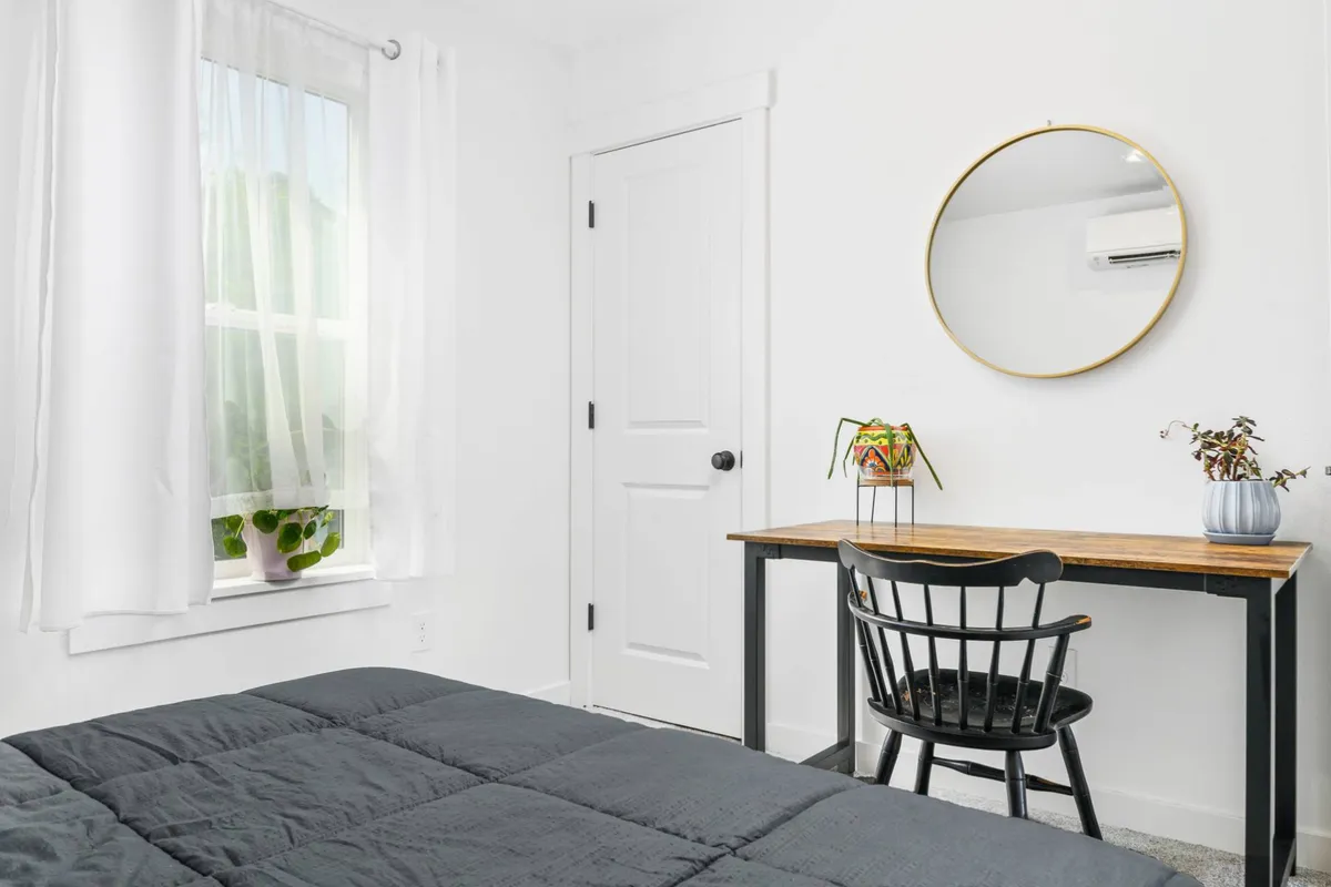 A serene, minimalistic space featuring a bed with dark bedding and a tidy desk by a window. The potted plant and soft light through sheer curtains create an inviting spot for reading, working, or relaxing, supporting focus and rest.