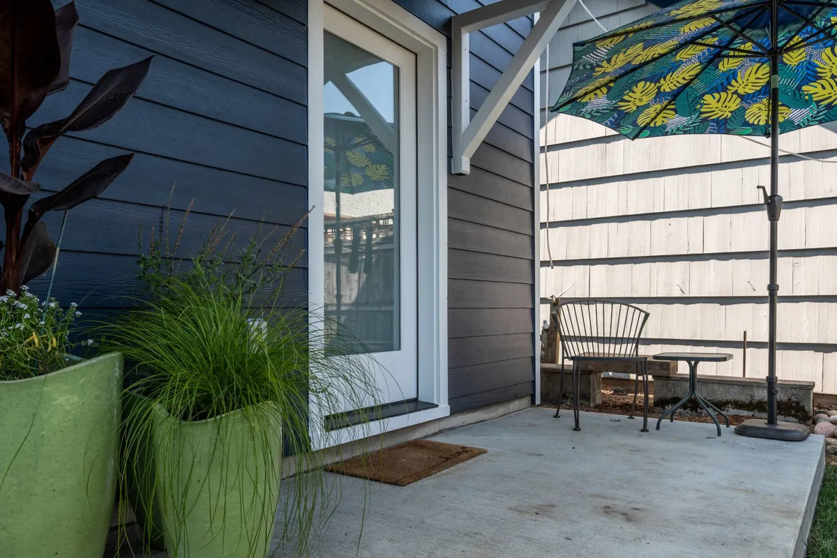 This serene exterior image displays a cozy outdoor seating area with green potted plants and a colorful umbrella. The space is perfect for relaxing with a book, enjoying morning coffee, or gathering with friends in a lush garden setting.