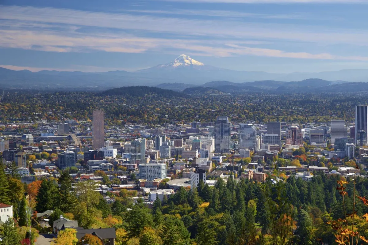A breathtaking view of the Portland city skyline with Mount Hood rising in the background and lush greenery in the foreground. The scene captures the accessibility of downtown Portland, scenic beauty, and opportunities for urban exploration.