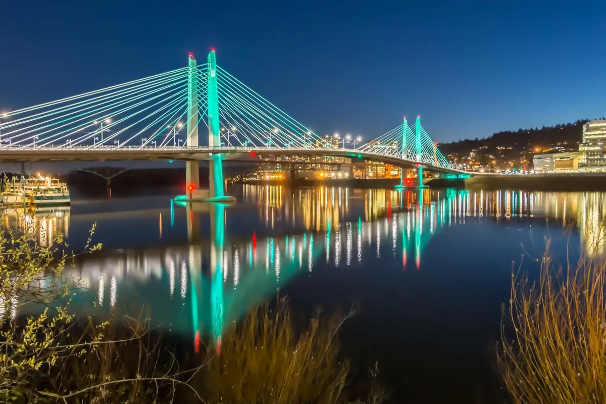 An atmospheric shot of an illuminated bridge at dusk, with city lights reflecting on the water and surrounding buildings in view. The scene evokes the tranquil yet lively spirit of the Portland waterfront, inviting guests to imagine evening strolls or riverside explorations close to the property.
