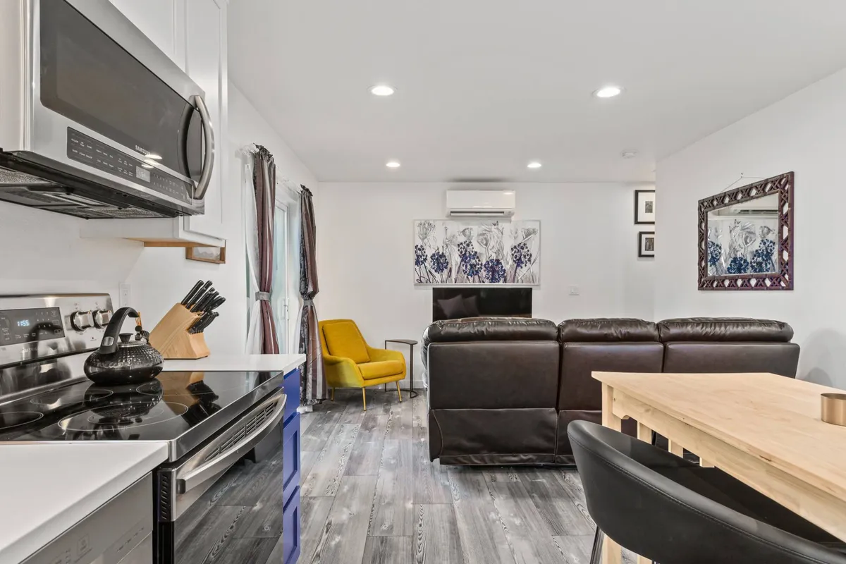 View of the open living area featuring a big brown sofa, yellow armchair, dining table, and visible kitchen appliances, all arranged for easy conversation and relaxation. The décor is contemporary with a cozy touch, suggesting a hub for entertaining and everyday living.