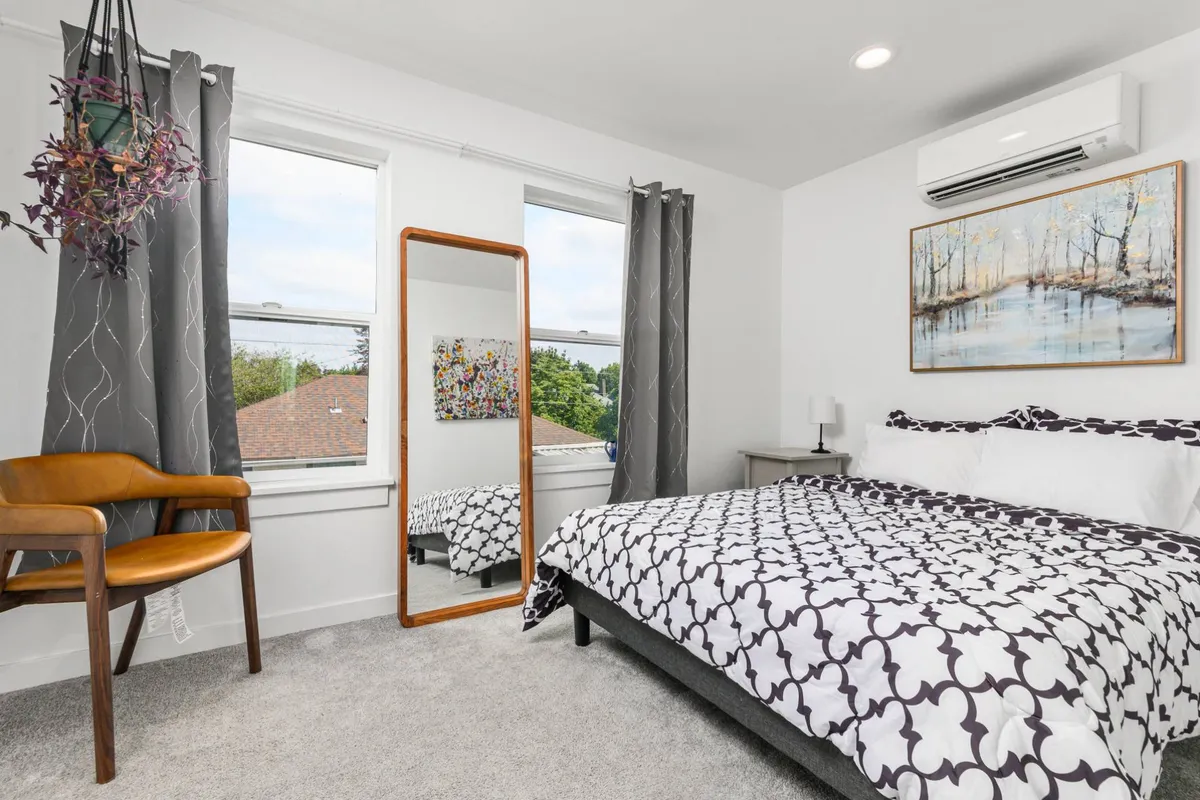A serene bedroom space with a comfortable bed, a decorative mirror, wall art, and a reading chair by the window. The layout and accessories suggest a personal, peaceful retreat ideal for unwinding, reading, or enjoying a quiet morning.